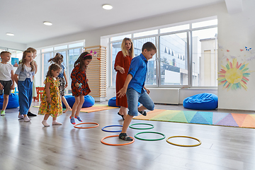 Image showing Small nursery school children with female teacher on floor indoors in classroom, doing exercise. Jumping over hula hoop circles track on the floor.