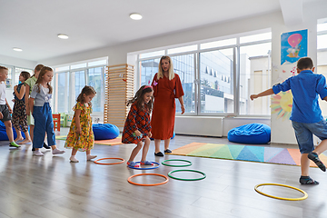 Image showing Small nursery school children with female teacher on floor indoors in classroom, doing exercise. Jumping over hula hoop circles track on the floor.