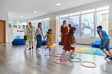 Image showing Small nursery school children with female teacher on floor indoors in classroom, doing exercise. Jumping over hula hoop circles track on the floor.