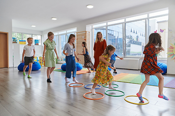 Image showing Small nursery school children with female teacher on floor indoors in classroom, doing exercise. Jumping over hula hoop circles track on the floor.