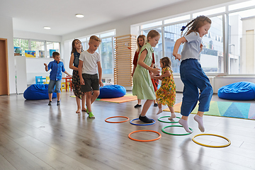 Image showing Small nursery school children with female teacher on floor indoors in classroom, doing exercise. Jumping over hula hoop circles track on the floor.