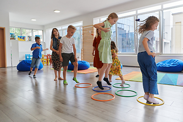 Image showing Small nursery school children with female teacher on floor indoors in classroom, doing exercise. Jumping over hula hoop circles track on the floor.