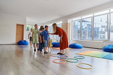 Image showing Small nursery school children with female teacher on floor indoors in classroom, doing exercise. Jumping over hula hoop circles track on the floor.