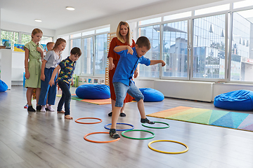 Image showing Small nursery school children with female teacher on floor indoors in classroom, doing exercise. Jumping over hula hoop circles track on the floor.