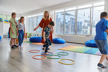 Image showing Small nursery school children with female teacher on floor indoors in classroom, doing exercise. Jumping over hula hoop circles track on the floor.