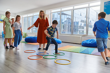 Image showing Small nursery school children with female teacher on floor indoors in classroom, doing exercise. Jumping over hula hoop circles track on the floor.