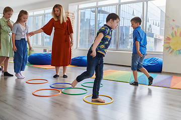 Image showing Small nursery school children with female teacher on floor indoors in classroom, doing exercise. Jumping over hula hoop circles track on the floor.