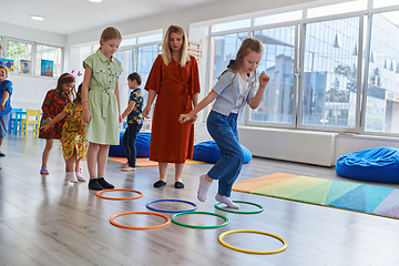 Image showing Small nursery school children with female teacher on floor indoors in classroom, doing exercise. Jumping over hula hoop circles track on the floor.