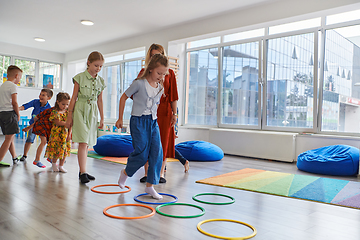 Image showing Small nursery school children with female teacher on floor indoors in classroom, doing exercise. Jumping over hula hoop circles track on the floor.