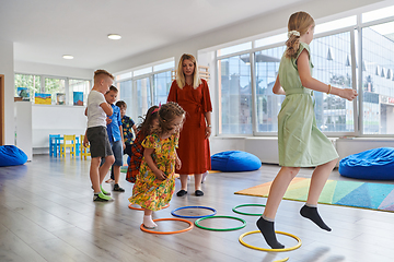 Image showing Small nursery school children with female teacher on floor indoors in classroom, doing exercise. Jumping over hula hoop circles track on the floor.