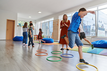 Image showing Small nursery school children with female teacher on floor indoors in classroom, doing exercise. Jumping over hula hoop circles track on the floor.