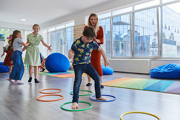 Image showing Small nursery school children with female teacher on floor indoors in classroom, doing exercise. Jumping over hula hoop circles track on the floor.