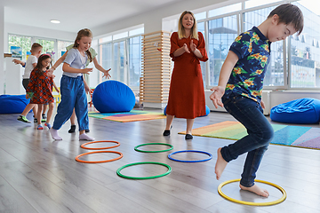 Image showing Small nursery school children with female teacher on floor indoors in classroom, doing exercise. Jumping over hula hoop circles track on the floor.