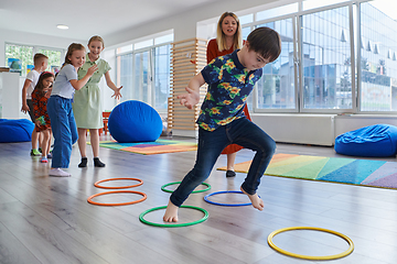 Image showing Small nursery school children with female teacher on floor indoors in classroom, doing exercise. Jumping over hula hoop circles track on the floor.