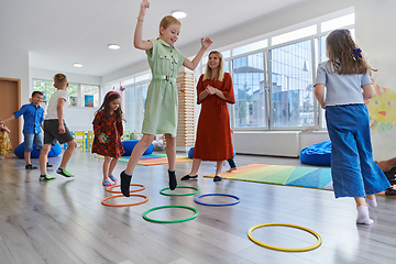 Image showing Small nursery school children with female teacher on floor indoors in classroom, doing exercise. Jumping over hula hoop circles track on the floor.