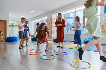 Image showing Small nursery school children with female teacher on floor indoors in classroom, doing exercise. Jumping over hula hoop circles track on the floor.