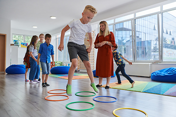 Image showing Small nursery school children with female teacher on floor indoors in classroom, doing exercise. Jumping over hula hoop circles track on the floor.