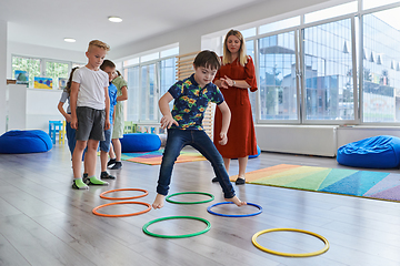 Image showing Small nursery school children with female teacher on floor indoors in classroom, doing exercise. Jumping over hula hoop circles track on the floor.