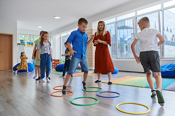 Image showing Small nursery school children with female teacher on floor indoors in classroom, doing exercise. Jumping over hula hoop circles track on the floor.