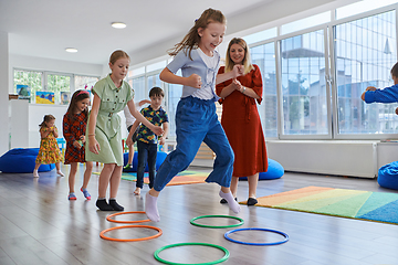 Image showing Small nursery school children with female teacher on floor indoors in classroom, doing exercise. Jumping over hula hoop circles track on the floor.