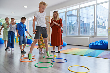 Image showing Small nursery school children with female teacher on floor indoors in classroom, doing exercise. Jumping over hula hoop circles track on the floor.