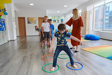 Image showing Small nursery school children with female teacher on floor indoors in classroom, doing exercise. Jumping over hula hoop circles track on the floor.