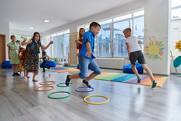 Image showing Small nursery school children with female teacher on floor indoors in classroom, doing exercise. Jumping over hula hoop circles track on the floor.