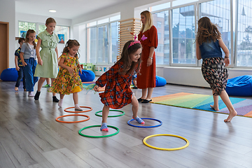 Image showing Small nursery school children with female teacher on floor indoors in classroom, doing exercise. Jumping over hula hoop circles track on the floor.