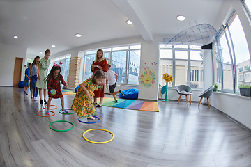 Image showing Small nursery school children with female teacher on floor indoors in classroom, doing exercise. Jumping over hula hoop circles track on the floor.