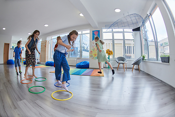 Image showing Small nursery school children with female teacher on floor indoors in classroom, doing exercise. Jumping over hula hoop circles track on the floor.