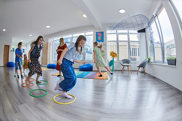 Image showing Small nursery school children with female teacher on floor indoors in classroom, doing exercise. Jumping over hula hoop circles track on the floor.