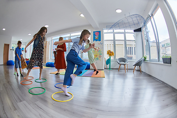 Image showing Small nursery school children with female teacher on floor indoors in classroom, doing exercise. Jumping over hula hoop circles track on the floor.