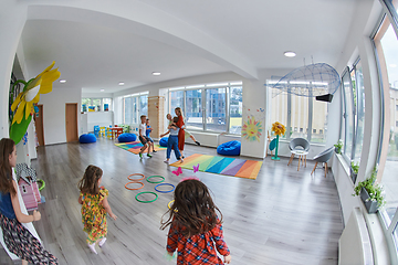 Image showing Small nursery school children with female teacher on floor indoors in classroom, doing exercise. Jumping over hula hoop circles track on the floor.