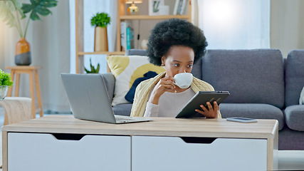 Image showing African American woman working remotely sitting on the floor having coffee with her tablet in her hand , laptop on coffee table and her back against the sofa in a living room