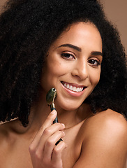 Image showing Face portrait, skincare and black woman with roller in studio on a brown background. Makeup, cosmetics and happy female model holding jade stone product for facial massage, healthy skin and beauty.