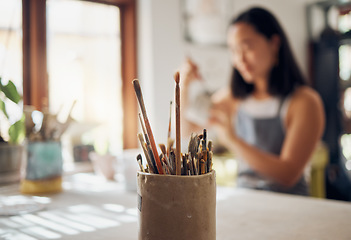 Image showing Paintbrush, art and equipment with a jar on a table in a pottery workshop or studio and a designer woman in the background. Zoom, paint and creative with a female potter working on a design