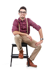 Image showing Nerd, smile and portrait of a man sitting on a chair wearing geek clothing and glasses. White background, happiness and smiling model on a stool in a studio looking positive, smart and hipster