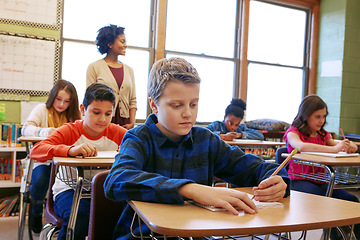 Image showing Boy child, classroom and writing at desk, school and focus on education, development and learning for future. School kids, teacher and studying at table in class with diversity, notebook and pencil