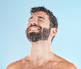 Image showing Man, shower and water for cleaning, studio and self care wellness with smile by blue background. Model, skincare and water splash on face, cold and self love for health, body and clean by backdrop