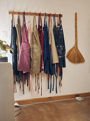 Image showing Clothes rack, apron and pottery with a broom in an empty workshop or studio against still life wall for design. Creative, art and clothing with a group of aprons hanging in a small business startup