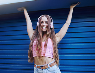 Image showing Portrait, music or freedom with a woman on a blue background standing arms raised in celebration for fun. Party, fashion and headphones with an attractive young female streaming or listening to audio