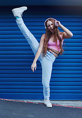 Image showing Crazy music, freedom and excited girl on blue background in city listening to track and audio on headphones. Fashion, lifestyle and woman with leg up for dance, happy attitude or streaming song