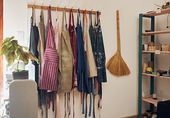 Image showing Clothes rack, apron and paint with a broom in an empty workshop or studio against still life wall for design. Creative, art and clothing with a group of aprons hanging in a small business startup