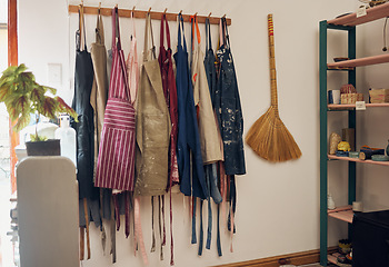 Image showing Apron, hanging and art shop with a broom in an empty workshop or studio against still life wall for design. Creative, pottery and clothing with a group of aprons on a rack in a small business startup