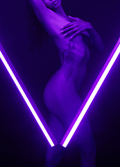 Image showing Nude, desirable and alluring woman posing naked in a studio. Beautiful, sensual and young woman behind purple lighting in a v shape. A female feeling sexy and covering her breasts in a dark studio