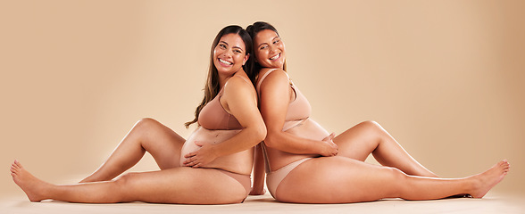 Image showing Pregnant woman friends, studio and underwear with smile, sitting and together for support by backdrop. Happy women, pregnancy and solidarity for wellness, care and holding healthy stomach by backdrop