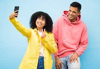 Image showing Friends, phone and peace sign for selfie on a blue background for fashion, style or friendship together. Young man and woman smiling and looking at smartphone for photo leaning on a wall in happiness