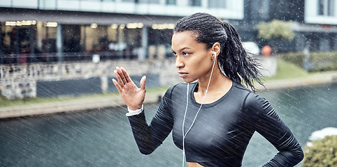 Image showing Black woman, fitness and running with earphones in the rain for sports motivation or determination in the city. African American sporty female runner doing intense cardio workout in the rainy weather