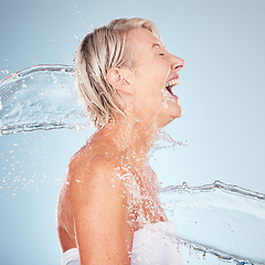 Image showing Woman, studio and happy with water splash, smile and cleaning for cosmetics by blue background. Excited senior model, skincare shower and cosmetic self care for health, aesthetic and body wellness