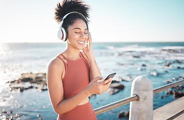Image showing Black woman at beach, headphone and smartphone with fitness, runner listening to music for sports motivation. Happy, streaming online with podcast or radio, phone and running by sea with calm outdoor